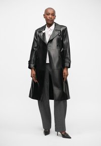 Schwarzer Leder-Trenchcoat mit doppelreihigen Knöpfen, ausgestattet mit einem taillierten Gürtel und breiten Revers, kombiniert mit grauen strukturierten Hosen.