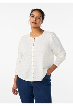 Moteris su garbanotais plaukais, dėvinti apskritas auskarus, tekstūruotą kreminės spalvos megztinio kardiganą su sagomis ir mėlynas džinsus, stovinti viena ranka kišenėje.