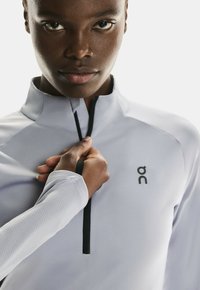 Helle graue Quarter-Zip-Oberbekleidung mit schwarzen Akzenten, gerippter Textur und einem Logo oben links. Die Hand des Modells justiert den Reißverschluss.