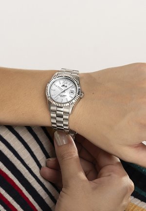 Silver metal wristwatch with white dial and date display worn on left wrist, being adjusted by right hand against striped clothing background.