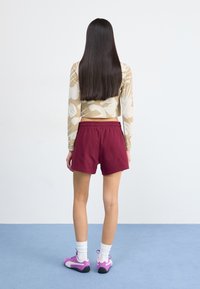 Haut à manches longues court avec un motif floral beige, associé à un short bordeaux et des chaussures de sport roses, vu de dos.