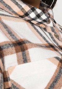Rutet flannel skjorte i hvid, tan og sort. Funktionen inkluderer en skjortekrave, frontknapper og en lomme med en spids flap.