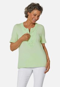 Top verde chiaro a maniche corte con scollatura testurizzata e ricamata. Tessuto liscio e vestibilità rilassata, abbinato a pantaloni bianchi.