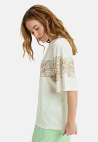 Cream cotton t-shirt with short sleeves, featuring a detailed brown floral design across the chest. Relaxed fit, soft texture.