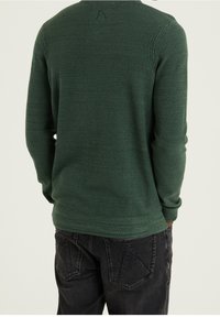 Grüner strukturierten Langarmpullover mit rundem Halsausschnitt und gerippten Akzenten, kombiniert mit dunklen Jeans mit einzigartigen Taschendesigns.