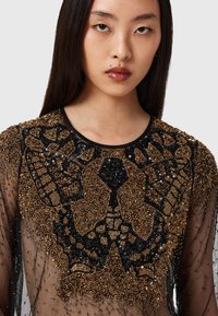 Schwarzes und golden verziertes Oberteil mit aufwendiger Perlenstickerei, einem Schmetterlingsmuster, transparenter Stoff und rundem Ausschnitt. Texturierte Details sind prominent hervorgehoben.