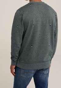 Person i en mörkgrå långärmad tröja med små abstrakta mönster, i kombination med blå denimjeans, sedd bakifrån.