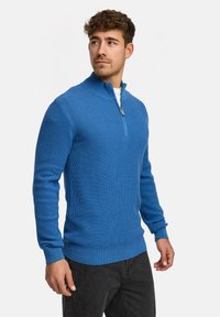 Blauer Strickpullover mit hohem Kragen und einem Viertel-Reißverschluss. Ausgestattet mit gerippten Texturen und Bündchen, kombiniert mit dunkelblauen Jeans.