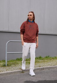 Brun sweatshirt med svart Nike-logotyp, vita sweatpants och vita sneakers. Modellen bär solglasögon och en blå skjorta under.