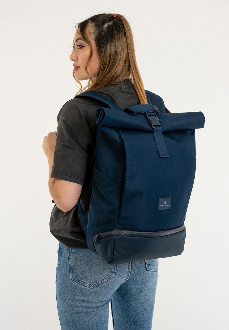 Jonge vrouw met een donkerblauwe rugzak met oprolbare bovenkant en ritspocket, gekleed in een zwart T-shirt en blauwe spijkerbroek, staand tegen een witte achtergrond.