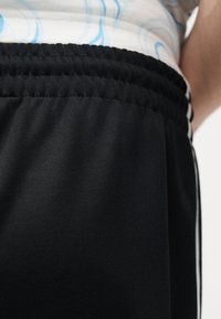 Κοντινό ενός ατόμου που φορά μαύρο παντελόνι με ελαστική μέση και λευκό πουκάμισο με ανοιχτό μπλε καμπυλωτές ρίγες.