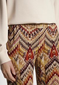 Pantalons à taille élastique avec un motif géométrique vibrant, présentant des rouges, des bruns et des jaunes sur un fond beige clair, fabriqués à partir d'un matériau doux.
