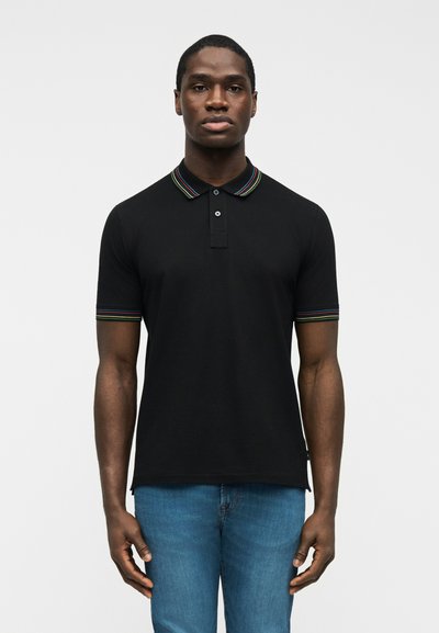 Homme portant un polo noir avec un col et des poignets de manches rayés multicolores, debout devant un fond blanc uni.