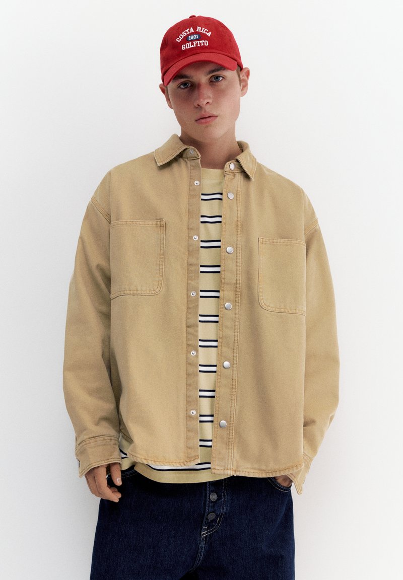Jeune homme portant une casquette rouge, une veste beige boutonnée sur une chemise rayée, et un jean bleu foncé, debout devant un fond blanc uni.