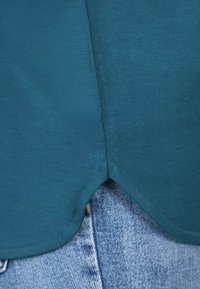 Türkisfarbene Stoffbluse mit abgerundetem Saum, aus glattem Material gefertigt. Sie überlappt leicht über einer hellblauen Jeans mit sichtbaren Nähten und Struktur.