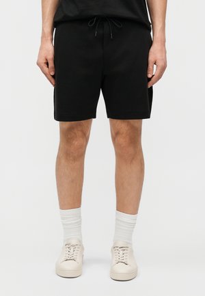 Homme portant un short noir à cordon, des chaussettes blanches montantes et des baskets beige à lacets, debout devant un fond clair uni.