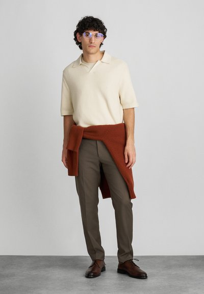 Kremna kratka majica polo iz pletenine, rjave hlače in rjavorjavi pleteni šal. Ima zaobljen ovratnik in očala s modrim odtenkom.