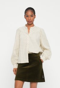 Cremefarvet ternet bluse med front bib-design, blødt stof og puffede lange ærmer; parret med en mørkegrøn corduroy mini-nederdel.