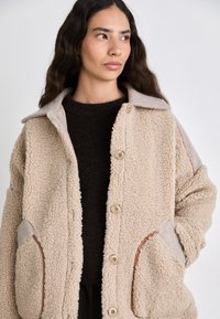 Giacca in pile beige con superficie testurizzata, dotata di colletto, quattro bottoni, due tasche laterali e dettagli grigi a contrasto.