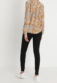 Koszula z długim rękawem w kolorze musztardowym z kwiatowym i zwierzęcym wzorem, zestawiona z czarnymi rurkami i białymi trampkami, widok z tyłu.