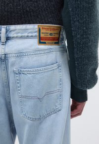 Light blue denim jeans with a visible back pocket, featuring a brown leather patch labeled "Diesel Industry Division." Textured dark sweater adjacent.