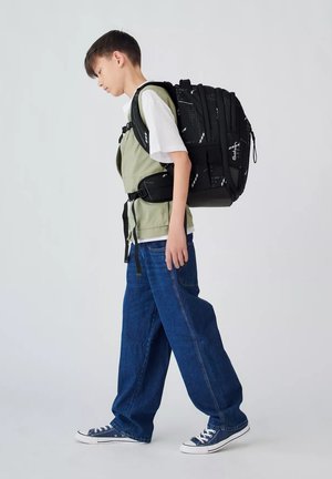 Schwarzer Rucksack mit gemusterten Akzenten, getragen von einem Kind in einer hellgrünen Weste, einem weißen T-Shirt und blauen weit geschnittenen Jeans. Blaue Sneakers.