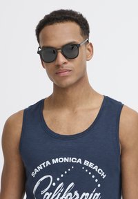 Črne sončne očala s okroglimi lečami, kovinski okvir, nosijo se s temno modrim majico brez rokavov, ki ima belo pisavo in okroglo grafično zasnovo.