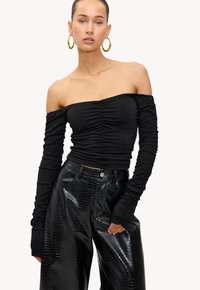 Vrouw in een geplooide zwarte off-shoulder top met lange mouwen en zwarte glanzende brede broek, vergezeld van grote gouden oorbellen.