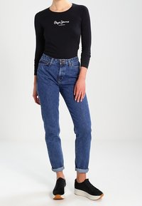 Schwarzes Langarmoberteil mit weißem Branding, kombiniert mit blauen Jeans mit hochgekrempelten Ärmeln und schwarzen Sneakern mit weißer Sohle und braunen Akzenten.