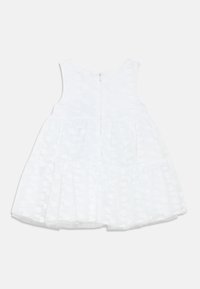 Robe blanche sans manches, avec un overlay texturé et une jupe à volants. Fermeture zippée au dos et détail en tissu à motif de cœurs.