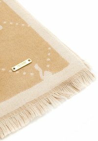 Beżowa i kremowa tkanina tkana z frędzlami oraz małą złotą metalką z napisem "GEHNIK" na białym tle.