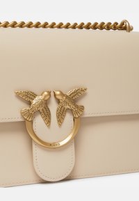 Borsa a tracolla in pelle sintetica beige con una catena dorata, caratterizzata da una chiusura a forma di due uccelli e un inserto metallico arrotondato.