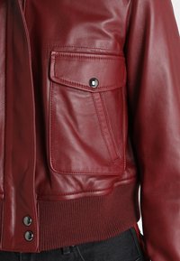 Röd läderbomberjacka med en framficka, ribbade ärmslut och knäppning med tryckknappar. Ytan är slät med en något glänsande finish.