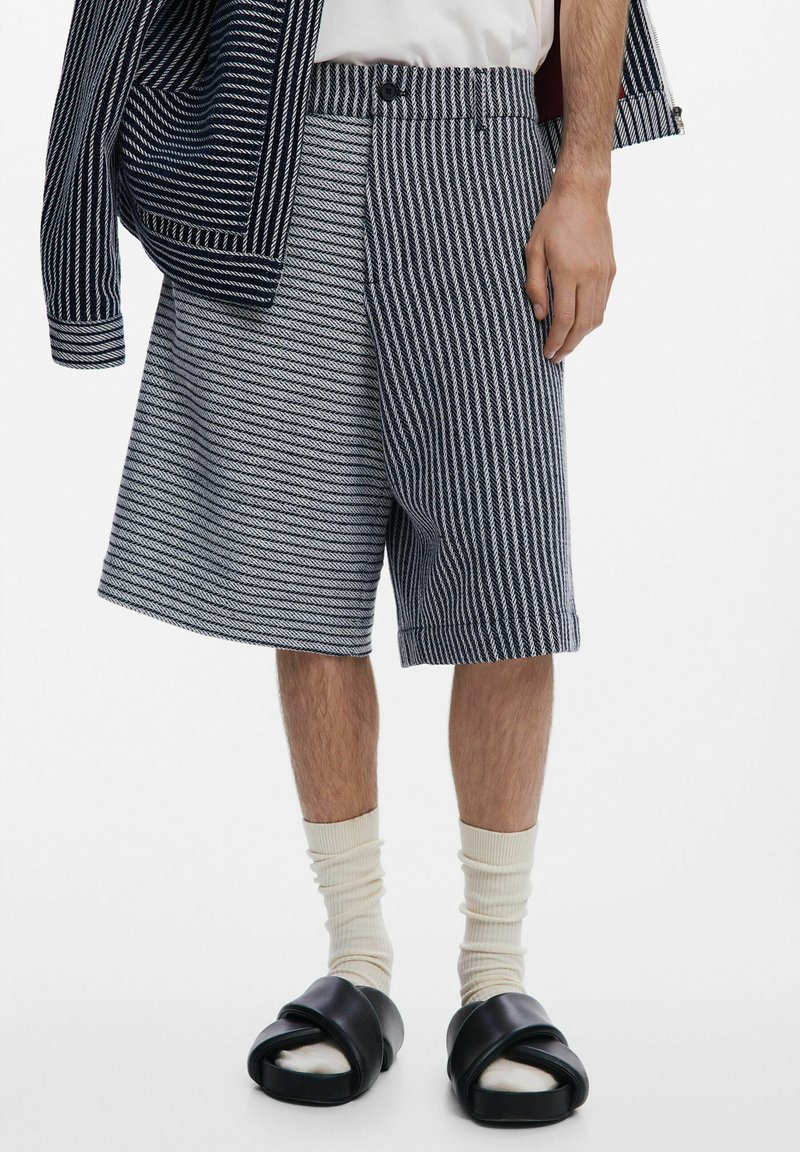 Csíkos, sötétkék és fehér anyagú rövidnadrág, laza fazonban, gombos derékkal és oldalsó zsebekkel, fekete szandálokkal párosítva.