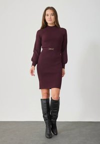 Abito in maglia color bordeaux con collo alto, trama a coste e maniche a sbuffo. Dotato di cintura con dettaglio dorato e stivali alti fino al ginocchio in pelle nera.