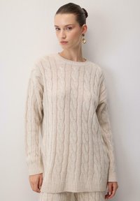 Beige neule, jossa on puikkoneulekuvio, pyöreä pääntie ja ribbaus hihansuissa. Pehmeä rakenne ja rento istuvuus, sopii kerroksittain käytettäväksi.