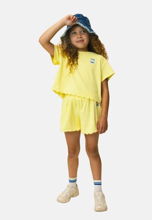 Kind mit lockigem Haar, das ein gelbes Outfit mit gewelltem Rand, weiße Turnschuhe, gestreifte Socken und einen blauen Eimerhut trägt und nach oben schaut.