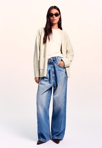 Cardigan oversized verde chiaro su una maglietta bianca, abbinato a jeans blu sbiaditi a gamba larga e occhiali da sole neri. Maglia a trama e denim liscio.