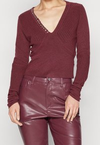 Bordowy sweter z prążkowanego dzianiny z głębokim dekoltem w kształcie litery V, ozdobiony srebrnymi elementami przy kołnierzu. Dopasowany krój i długie rękawy. W komplecie z pasującymi spodniami.