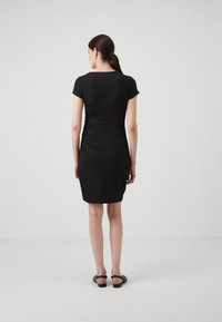Robe noire à manches courtes avec une silhouette cintrée, des détails froncés sur les côtés et un tissu lisse et extensible. Portée avec des sandales noires.