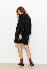 Robe noire avec des manches bouffantes et des détails brodés. Présente une coupe ample et une longueur au genou, associée à des chaussettes texturées rouges et des mocassins.