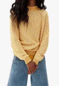 Personne aux longs cheveux bouclés portant un pull jaune en tricot texturé et un jean bleu, debout les mains légèrement jointes devant.