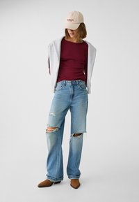Bordeaux rood lange mouwen top, grijze sweatshirt gedrapeerd, lichtblauwe versleten spijkerbroek, bruine puntige laarzen, beige pet met rode tekst.
