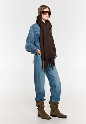 Giacca di jeans, jeans a gamba ampia blu, sciarpa frangiata marrone, stivaletti con tacco marroni, occhiali da sole neri grandi, sfondo neutro.