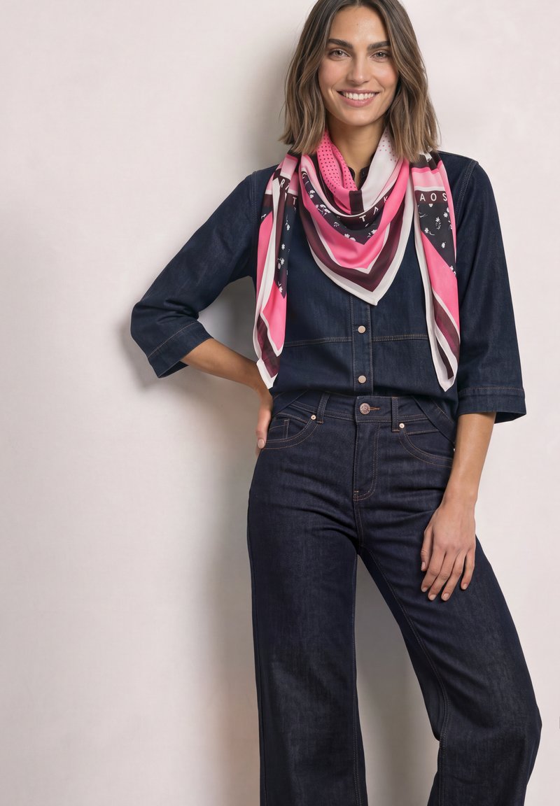 Femme en tenue en jean bleu foncé souriante, debout avec une main sur la hanche, portant une grande écharpe rose, blanche et noire à motifs autour du cou.