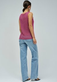 Magenta pleteni top bez rukava s teksturiranim uzorkom, u kombinaciji s širokim svijetloplavim trapericama i crnim sandalama s potpeticama. Pogled straga.