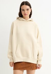 Sudadera con capucha beige con un gran bolsillo frontal, puños acanalados y un ajuste cómodo. La tela parece suave con una textura lisa, combinada con una falda marrón estampada.