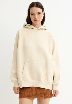 Sudadera con capucha beige con un gran bolsillo frontal, puños acanalados y un ajuste cómodo. La tela parece suave con una textura lisa, combinada con una falda marrón estampada.