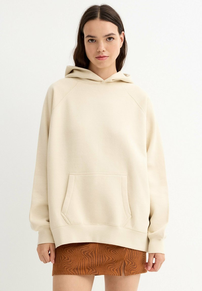Sudadera con capucha beige con un gran bolsillo frontal, puños acanalados y un ajuste cómodo. La tela parece suave con una textura lisa, combinada con una falda marrón estampada.