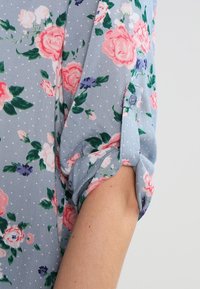 Blusa floreale con sfondo azzurro chiaro, rosa e violette, pois, maniche arrotolate e dettaglio con bottone sui polsini.